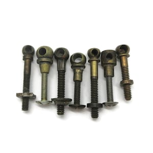 May include: A collection of seven vintage-style metal bolts with eyelet heads, each featuring a threaded shaft and a distinct brass or bronze-toned finish. These hardware components vary slightly in shape and size, displaying a weathered patina.