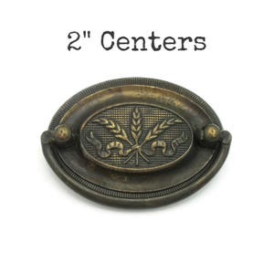 May include: An oval-shaped brass drawer pull with a dark, aged finish. The center features a relief design of wheat stalks tied with a ribbon against a grid background. The hardware includes a swinging handle and is labeled 2" Centers.