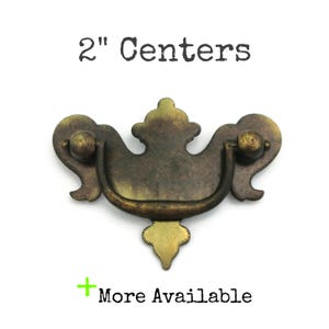 May include: A 2" center antique brass cabinet pull with a decorative, ornate design. The pull has a curved handle and a decorative, ornate design.