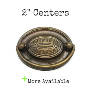 May include: Antique brass drawer pull with a decorative oval center and a handle. The text "2" Centers" is above the pull, and "More Available" is below. The pull measures 5.08 cm.