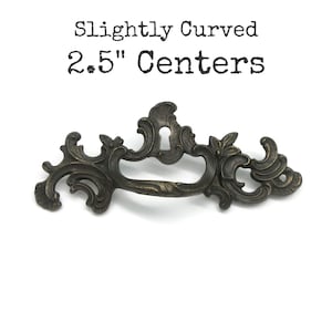 May include: A dark bronze-toned metal drawer pull with an ornate, scrolling design and a central keyhole cutout. The hardware features a swinging handle and is labelled with the text Slightly Curved 2.5" Centers above the object.