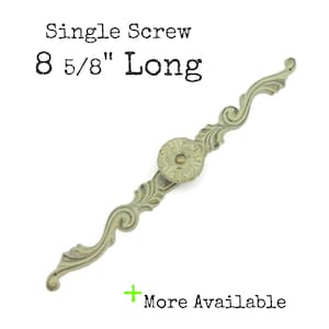 May include: An antique-style, decorative cabinet pull with a single screw. The pull is 8 5/8 inches long and features an ornate, scrolling design with a central circular element. The finish is a muted, aged green with hints of gold.