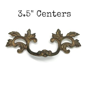 May include: A bronze-toned metal drawer pull featuring an ornate, symmetrical scrollwork design with leaf-like motifs. The hardware has a dark, weathered finish and is positioned below the black text that reads 3.5" Centers.