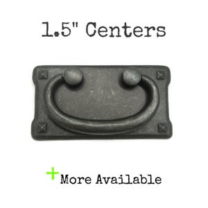 May include: A dark gray metal drawer pull with a rectangular backplate featuring four small pyramid-shaped studs at the corners and a curved drop handle. The text 1.5" Centers and + More Available is printed above and below the hardware.