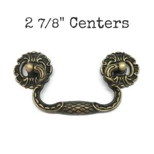 May include: A brass-toned metal drawer pull with a drop handle featuring a textured diamond pattern in the center and ornate circular backplates. The text 2 7/8" Centers is printed above the hardware, indicating the distance between the mounting points.
