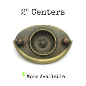 May include: Antique brass-toned drawer pull with a central circular design and two end knobs. The text "2" Centers" is above the pull, and "More Available" is below. The pull measures 5.08 cm across.