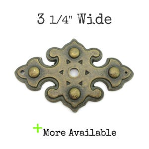 May include: Antique brass-toned decorative furniture hardware, 3 1/4" wide, with a central hole and four rounded accents. The hardware has a detailed, ornate design with a vintage aesthetic. The text "More Available" is visible.