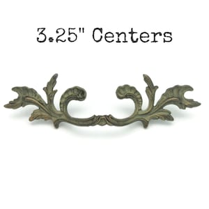May include: An antique brass-toned drawer pull with an ornate, floral design. The pull has a 3.25-inch center-to-center measurement. The decorative hardware features a curved shape with detailed leaf and scroll elements, perfect for vintage furniture restoration.