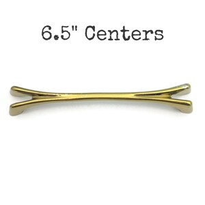 May include: A gold-toned cabinet pull with a distinctive bow-tie shape. The text "6.5" Centers" is printed above the pull. The pull measures 6.5 inches across.