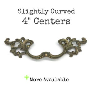 May include: An antique brass-toned metal drawer pull featuring ornate, symmetrical scrollwork and leaf-like detailing. The text above reads Slightly Curved 4" Centers, and the text below reads + More Available.
