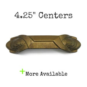 May include: A brass-toned metal drawer pull with a central rectangular bar and decorative fan-shaped ends. The text 4.25" Centers is printed above the object, and a green plus sign with the text More Available is printed below.