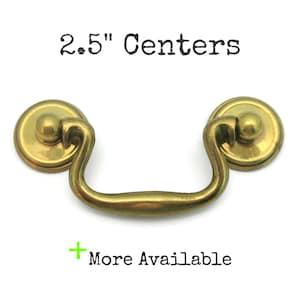 May include: A brass-toned drawer pull with a curved handle and two circular backplates. The text "2.5" Centers" is above the pull, and "+ More Available" is below. The pull measures 2.5 inches between the centers of the mounting holes.