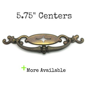 May include: An antique-style drawer pull with a bronze finish. The pull has an oval center with a decorative keyhole design and a curved handle. The text "5.75" Centers" is above the pull, and "More Available" is below.