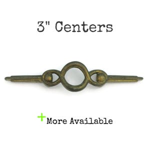May include: A bronze-colored cabinet pull with a central ring design and two decorative end pieces. The text "3" Centers" is above the pull, and "More Available" is below. The pull measures 3 inches in length.