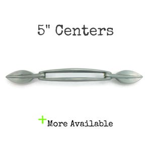 May include: A silver-toned cabinet pull with a 5-inch (12.7 cm) center-to-center measurement. The pull has a slender, curved handle and decorative, leaf-shaped ends. The text "5" Centers" and "More Available" are also visible.