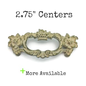 May include: Antique-style drawer pull with a floral design. The pull is approximately 2.75 inches (7 cm) between the centers of the mounting holes. The metal has a distressed, aged appearance with a cream and gold finish. The text "2.75" Centers" is visible.