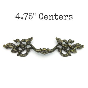May include: Antique brass drawer pull with ornate floral detailing. The pull measures 4.75 inches between the centers of the mounting holes. The handle has a curved design and is suitable for cabinets and drawers.