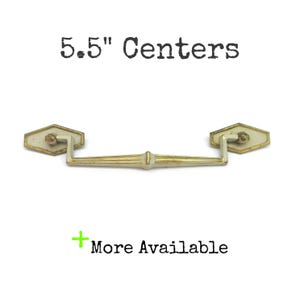 May include: A metal drawer pull with a gold and off-white finish, featuring a central bar and two hexagonal mounting plates. The text 5.5" Centers and + More Available is visible. The hardware has a weathered appearance with visible gold trim.