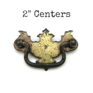 May include: Antique brass drawer pull with a decorative design. The pull has a curved handle and measures 5.08 cm between the centres of the mounting holes. The text "2" Centers" is above the pull.