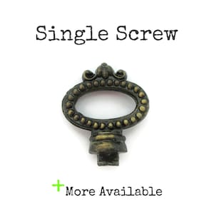 May include: A bronze-toned metal furniture knob with an oval shape and a beaded border. The top features a decorative crest. The text "Single Screw" appears above the knob, and the text "+ More Available" is printed below the object.