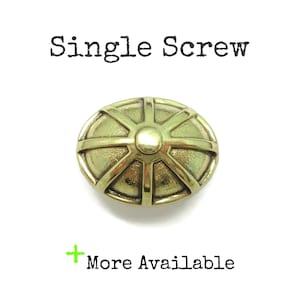 May include: A brass-toned oval knob featuring a raised sunburst pattern with a central circular dome. The text "Single Screw" appears above the knob, and the text "+ More Available" is positioned below the object on a plain white background.