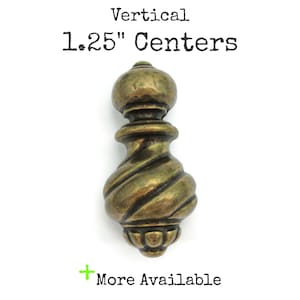 May include: Antique brass-finished cabinet pull with a vertical design. The pull features a decorative, spiraled body and measures 1.25 inches in center-to-center distance. The text "More Available" is also visible.