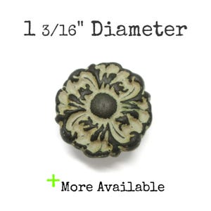 May include: A round, floral-shaped knob featuring a dark brown and off-white distressed finish. The text 1 3/16" Diameter is printed above the knob, and the text + More Available is printed below the knob on a plain white background.