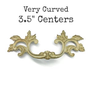 May include: A curved, off-white drawer pull with a distressed finish and ornate leaf-like detailing. The text "Very Curved 3.5" Centers" is printed above the hardware, indicating the distance between the mounting points in inches.
