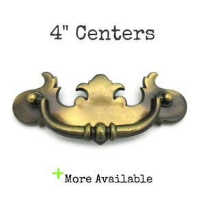 May include: Antique brass drawer pull with a decorative design. The pull has a curved handle and measures 4 inches (10.16 cm) between the centers of the mounting holes. The text "4" Centers" is above the pull, and "More Available" is below.