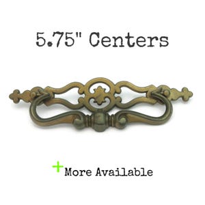 May include: Antique brass drawer pull with a decorative design. The pull has a curved handle and ornate detailing. The text "5.75" Centers" is above the pull, and "More Available" is below it.