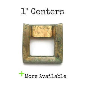 May include: A square, antique brass cabinet pull with a 2.54 cm centre-to-centre measurement. The hardware has a rectangular shape with a central opening. The text "1" Centers" is above the pull, and "More Available" is below.