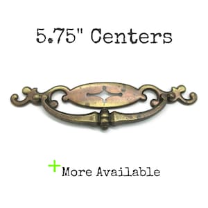 May include: A brass-toned metal drawer pull with an oval backplate and a curved handle. The text 5.75" Centers is displayed above the hardware, and the text + More Available is positioned below the object.