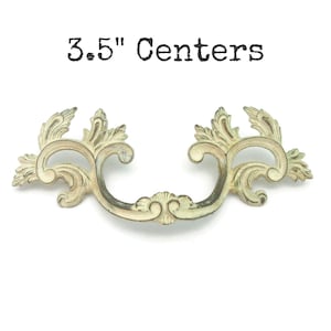 May include: Ornate antique-style drawer pull with a cream and aged bronze finish. The handle is 3.5 inches (8.9 cm) between the screw centers, featuring a scrolling leaf design and a central decorative element. The text "3.5" Centers" is above the handle.