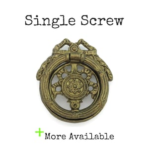 May include: Antique brass-toned drawer pull with a ring handle and decorative floral design. The text "Single Screw" is above the pull, and "More Available" is below. The pull is centered on a white background.