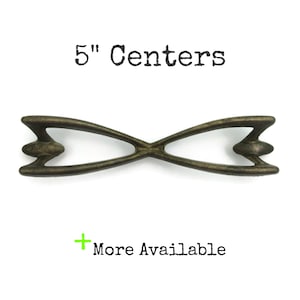 May include: A bronze-toned metal cabinet pull with an X-shaped design and pointed ends. The text 5" Centers is printed above the pull, and the text + More Available is printed below the pull in black lettering on a plain white background.