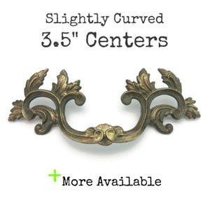 May include: Antique brass-toned drawer pull with a curved design. The pull measures 3.5 inches between the centers of the mounting holes. The handle features ornate, swirling leaf and scroll details, and the text "Slightly Curved 3.5" Centers" is visible.