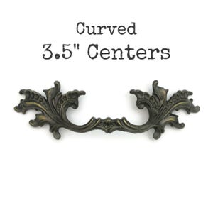 May include: An antique-style drawer pull with a curved design and ornate floral detailing. The bronze-colored handle has a 3.5-inch (8.89 cm) center-to-center measurement. The text "Curved 3.5" Centers" is printed above the handle.