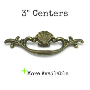 May include: An antique brass-coloured metal drawer pull with a central shell motif and curved arms. The text 3" Centers is printed above the handle, and the text + More Available is printed below the handle on a plain white background.