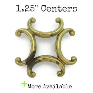 May include: A brass decorative hardware piece with a square centre and curved, scroll-like arms. The text "1.25" Centers" is at the top, and "More Available" is at the bottom. The hardware has a tarnished appearance.