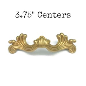 May include: A gold-toned metal drawer pull with an ornate, curved design and scrolling details. The text 3.75" Centers is printed above the hardware, indicating the distance between the mounting holes for this specific furniture handle.