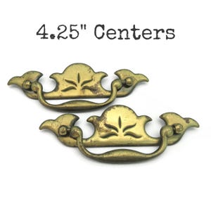 May include: Two antique brass drawer pulls with a distressed finish. Each pull features a decorative floral design and a curved handle. The text "4.25" Centers" is printed above the pulls.
