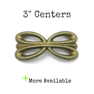 May include: A brass-toned metal drawer pull with a symmetrical, looped, bow-like design. The hardware features a smooth, matte finish and an open center. The text 3" Centers is printed above the object, and the text + More Available is below.