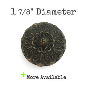 May include: A bronze-colored, round cabinet knob with a floral design. The knob is 1 7/8" in diameter and features a central flower surrounded by detailed leaf patterns. The text "More Available" is below the knob.