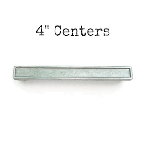 Peut inclure: Une poignée de meuble rectangulaire argentée avec un centre encastré. Le texte "4" Centers" est imprimé au-dessus de la poignée. La poignée mesure 10,16 cm de long.