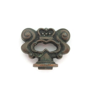 May include: A dark bronze-toned metal key bow featuring an ornate, symmetrical design with scrollwork accents and a central crown motif. The piece has a weathered, aged patina with visible dark brown and subtle green oxidation across the surface.