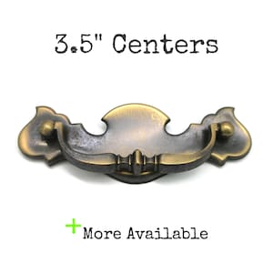May include: Antique brass drawer pull with a decorative design. The pull has a curved handle and measures 3.5 inches between the centers of the mounting holes. The text "3.5" Centers" is above the pull, and "More Available" is below.