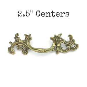 May include: Ornate brass drawer pull with a curved handle and decorative floral accents. The text "2.5" Centers" is above the pull. The pull measures 6.35 cm between the mounting holes, suitable for furniture restoration or DIY projects.