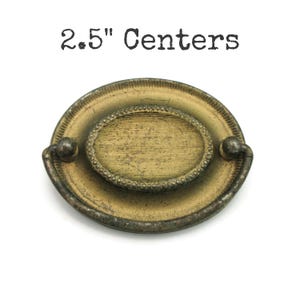 May include: An aged brass drawer pull with an oval centre and beaded trim. The pull has a 6.35 cm centre-to-centre measurement and features a distressed finish, giving it a vintage appearance.
