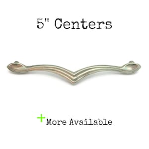 May include: A silver-toned cabinet pull with a V-shaped design, measuring 5 inches between the centers of the mounting holes. The text "5" Centers" is above the pull, and "More Available" is below.