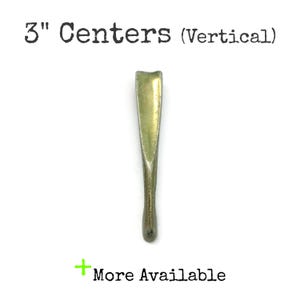 May include: A vertical brass-toned metal cabinet pull with a smooth, tapered design. The text 3" Centers (Vertical) is printed above the handle, and a green plus sign with the text More Available is printed below the handle.